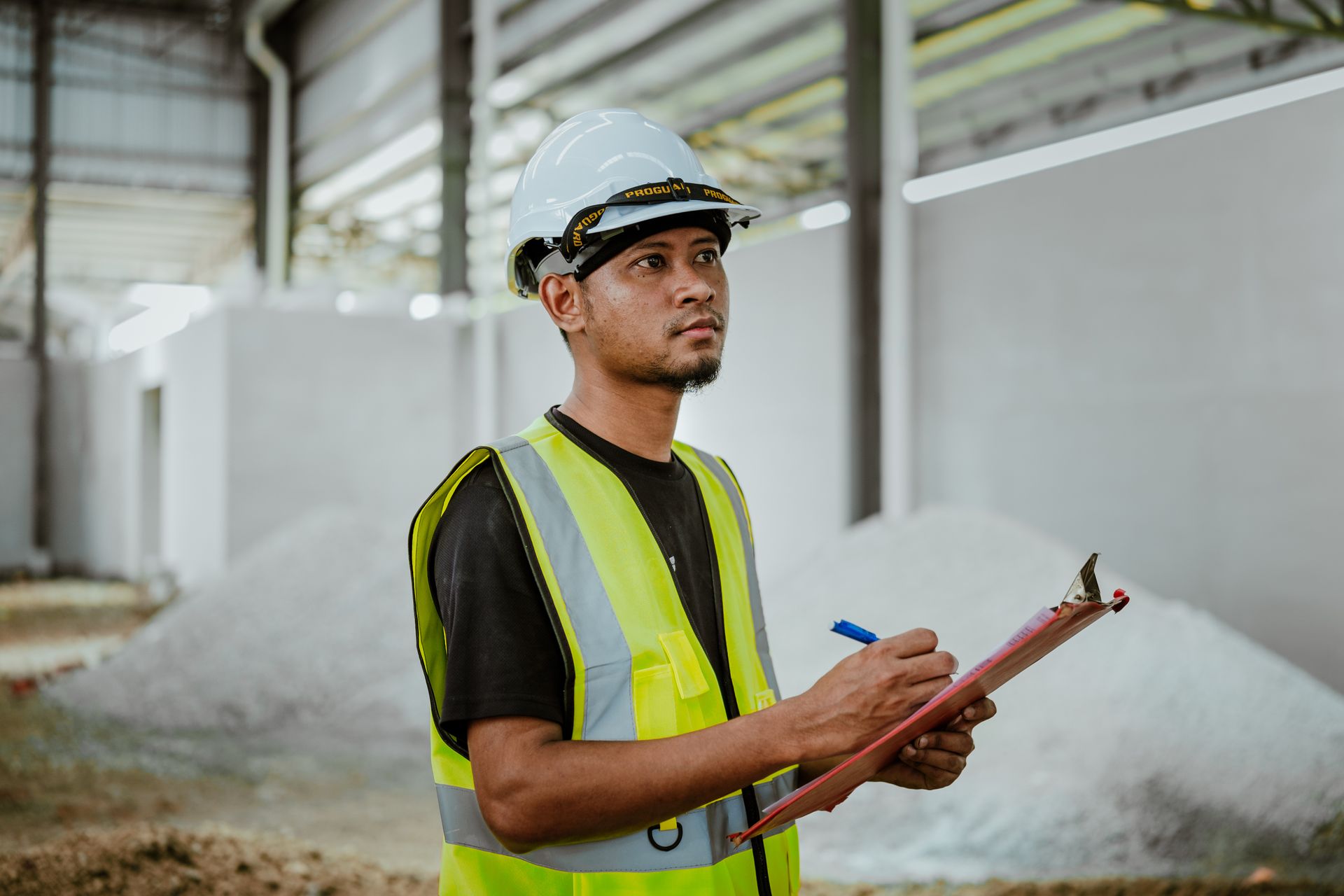 Worker in a hard hat and high-visibility vest reviewing notes on a clipboard inside an industrial plant